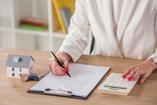 Partial-view-of-businesswoman-using-calculator-and-writing-in-contract-near-house-model-and-keys